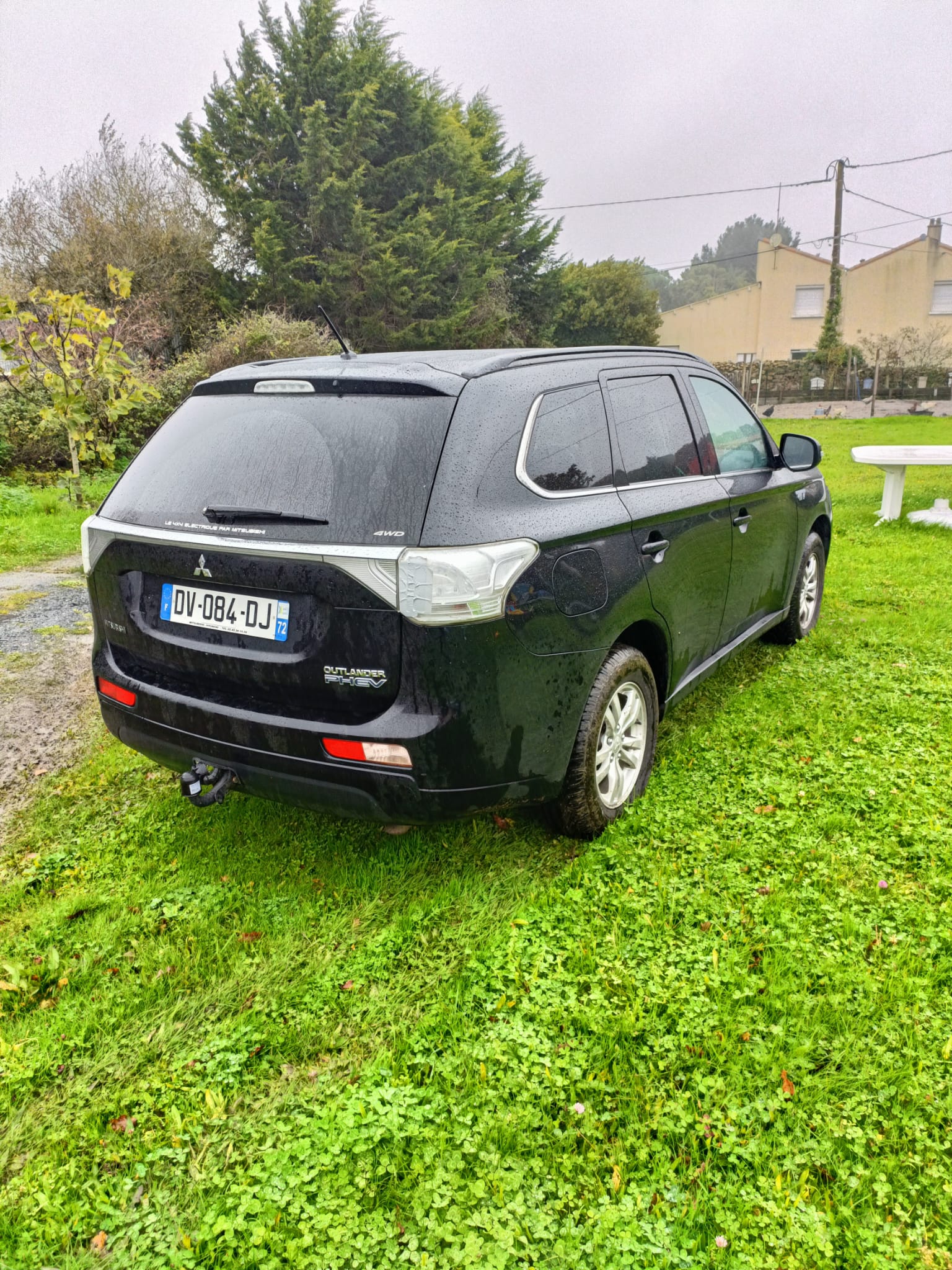 Mitsubishi Outlander PHEV noir vu de trois-quarts arrière, garé sur une pelouse avec attelage et arbres derrière